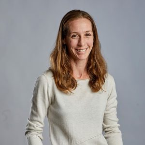 A white woman with sandy brown hair stands against a blank background and smiles professionally at the camera.