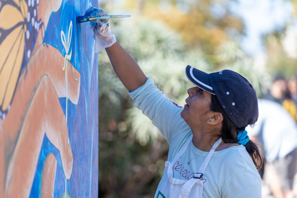 A dark-skinned woman in a navy blue ballcap reaches up with a gloved hand to add more blue paint to a mural of a monarch butterfly on a dark-skinned hand. Her dark hair is pulled back out of her face while she works, and her white shirt and apron are unmarked by paint.