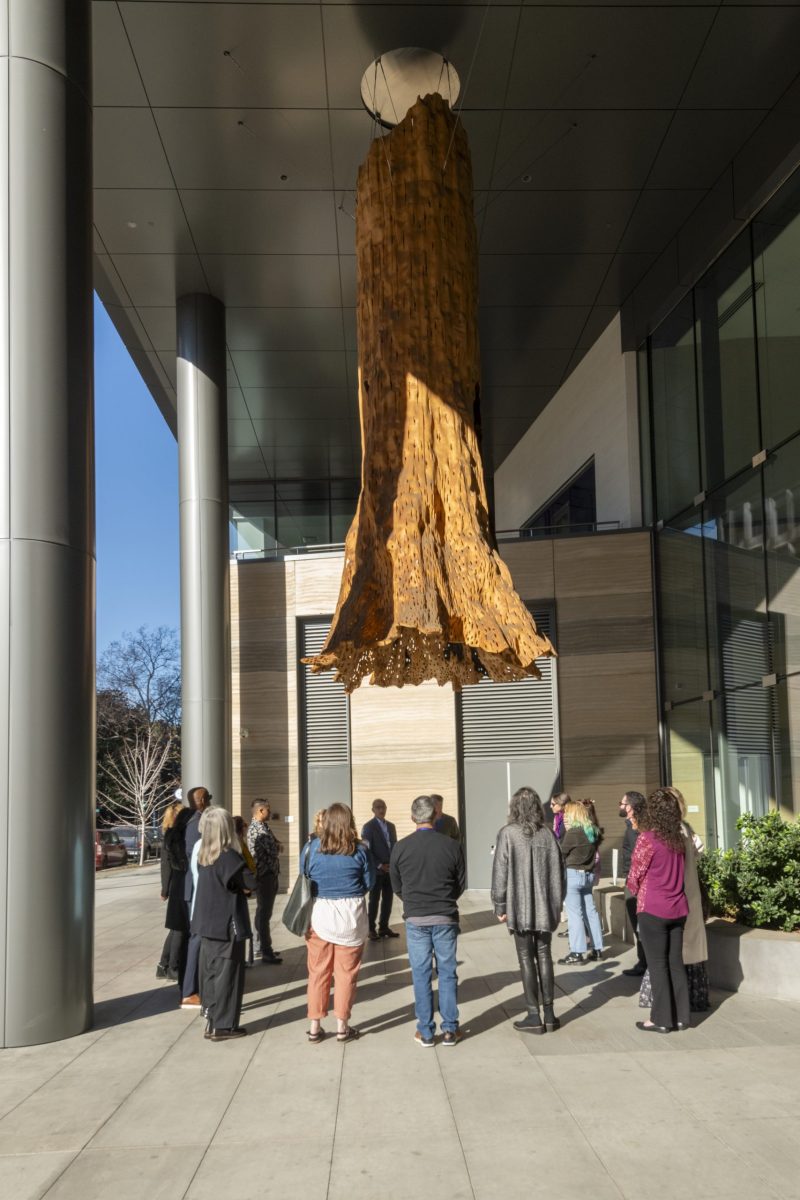A group of people stand in a circle just beyond the reach of a hanging sculpture. It's outdoors and the sunshine is streaming in at an angle across the bottom half. The sculpture looks like a wilted yellow trumpet flower, and is hanging from the roof overhang several stories overhead.