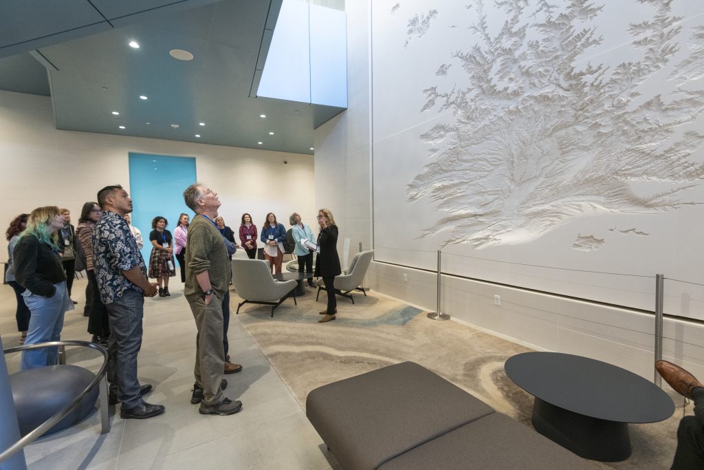 A group of people are dwarfed by the large artwork they're looking at. It's a two-story high white wall with a relief map carved into it representing what looks like a river basin.
