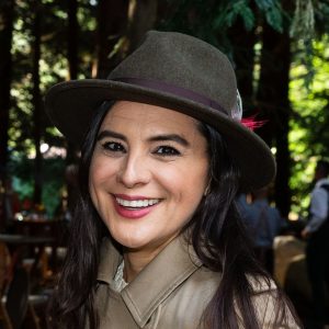 A woman whose dark hair blends into the trees behind wears a felt hat with a narrow brim and smiles at the camera.