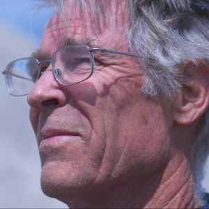 An older white man with glasses and grey hair looks off into the sunshine.