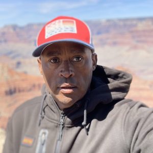 A brown-skinned man in a baseball cap that's shading his face from the bright sun is taking a selfie in a dry, arid place.