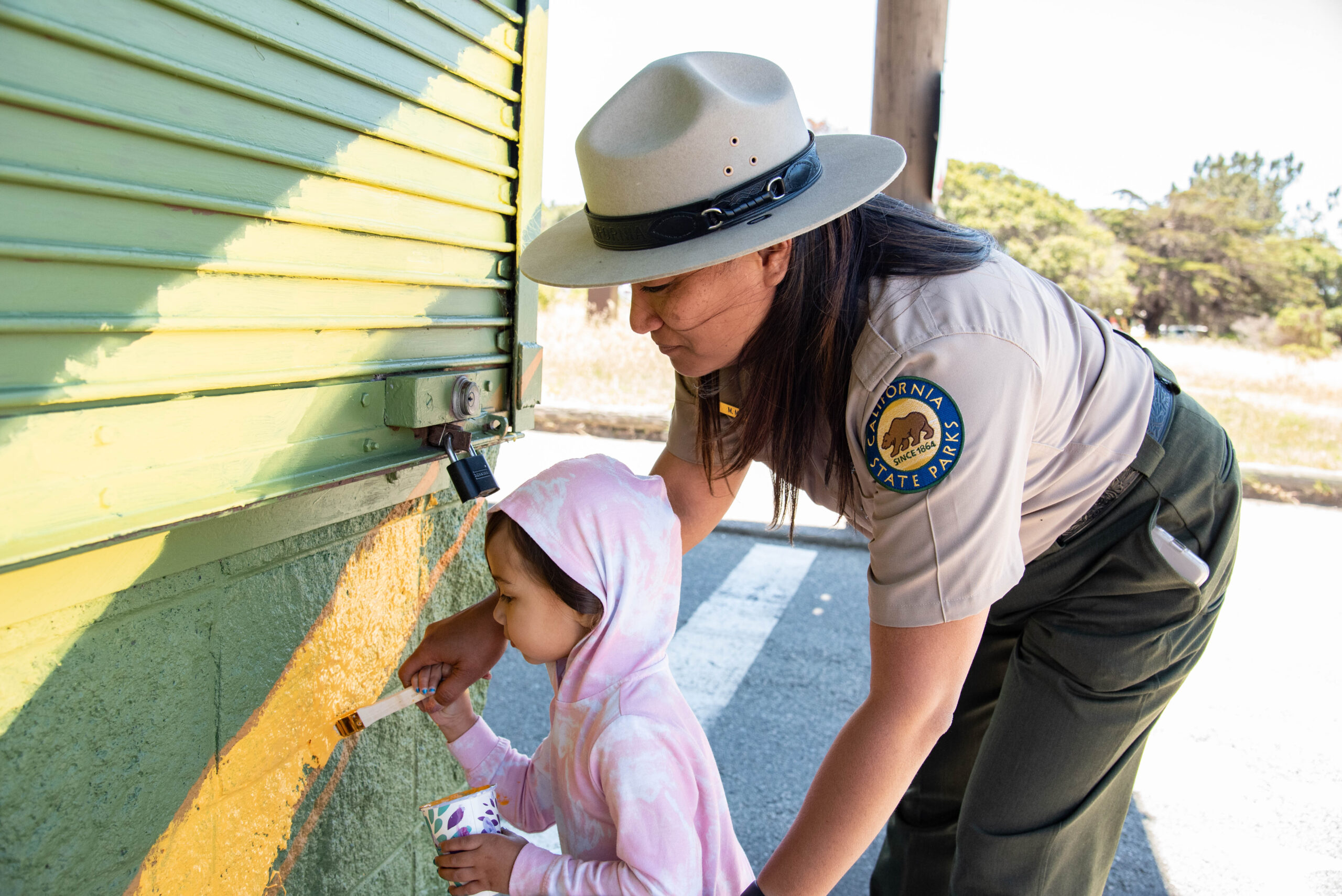A park ranger in uniform with long dark hair bends down to help a small child in a pink hoodie carefully brush yellow paint along the side of a building as part of a mural. Several stripes of other muted earthy colors already adorn the concrete and plastic siding, looking like bright streaks of sunshine.