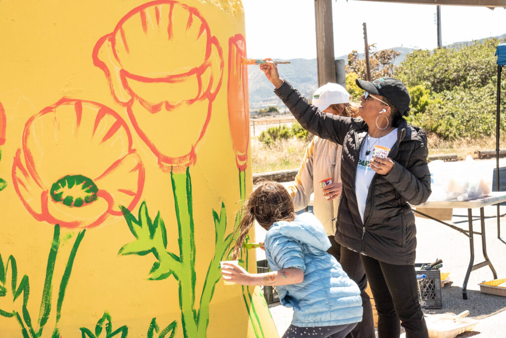 Two adults reach up to continue painting California poppy flowers on a yellow mural wall. The poppies and their leaves are outlined, and the leaves are being colored in by a smaller person reaching in front of the adults. It looks like a fun, collaborative activity.