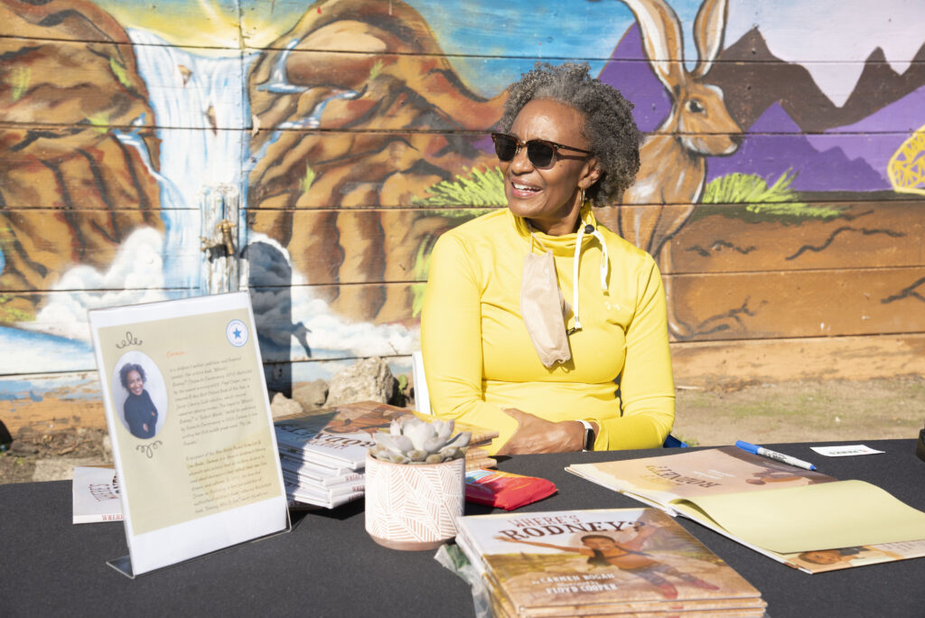 A Black femm in a yellow cardigan and stylish sunglasses sits behind a table displaying the book she wrote. She is looking to the left and smiling at someone off camera. The bright sunshine makes her glow with happiness. Behind her is a fanciful but semi-realistic mural depicting a waterfall in a desert and a hare that seems to be looking over Carmen's shoulder.