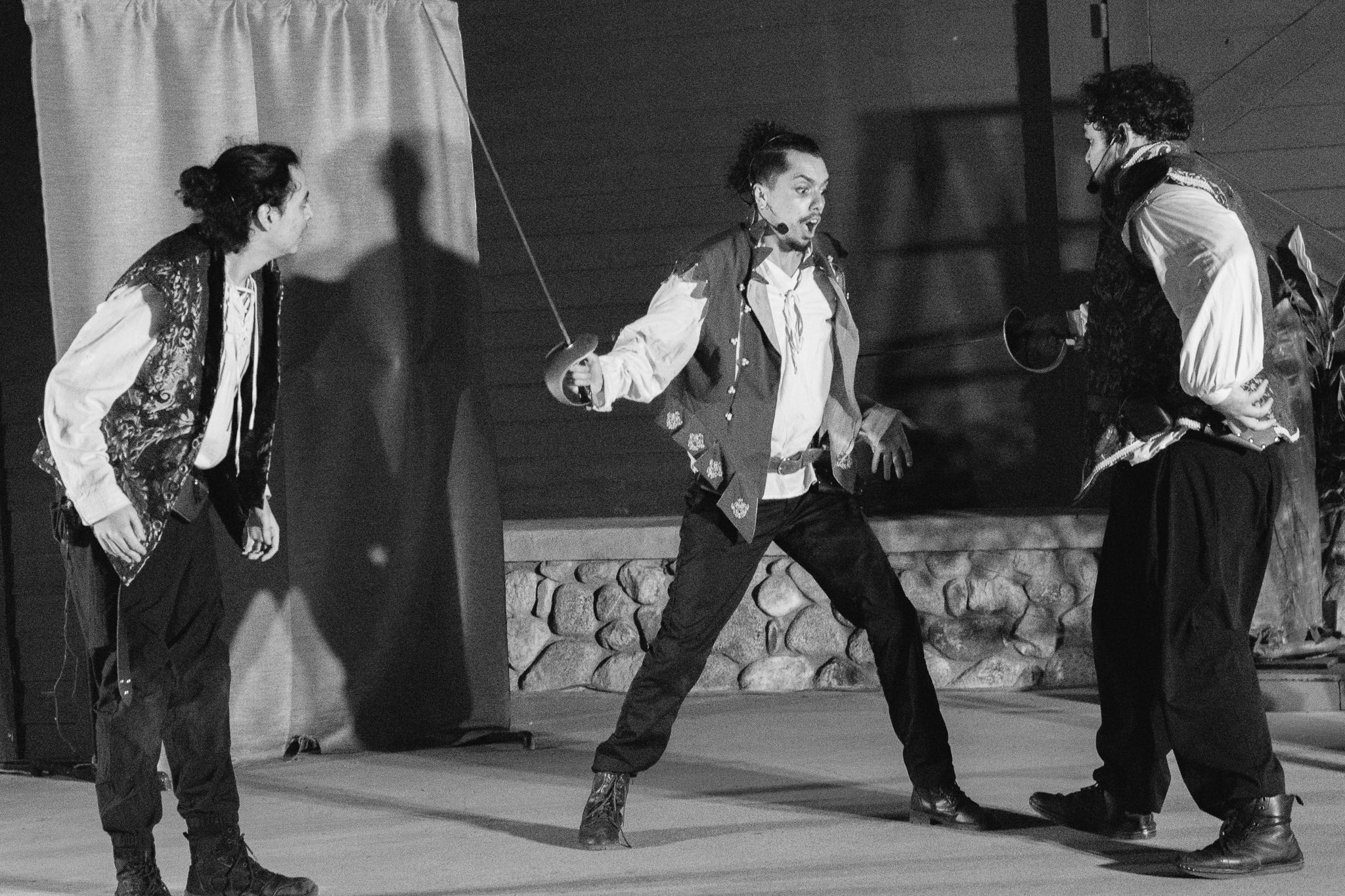 Three young men sword fight with fencing foils, dressed in Renaissance costumes.