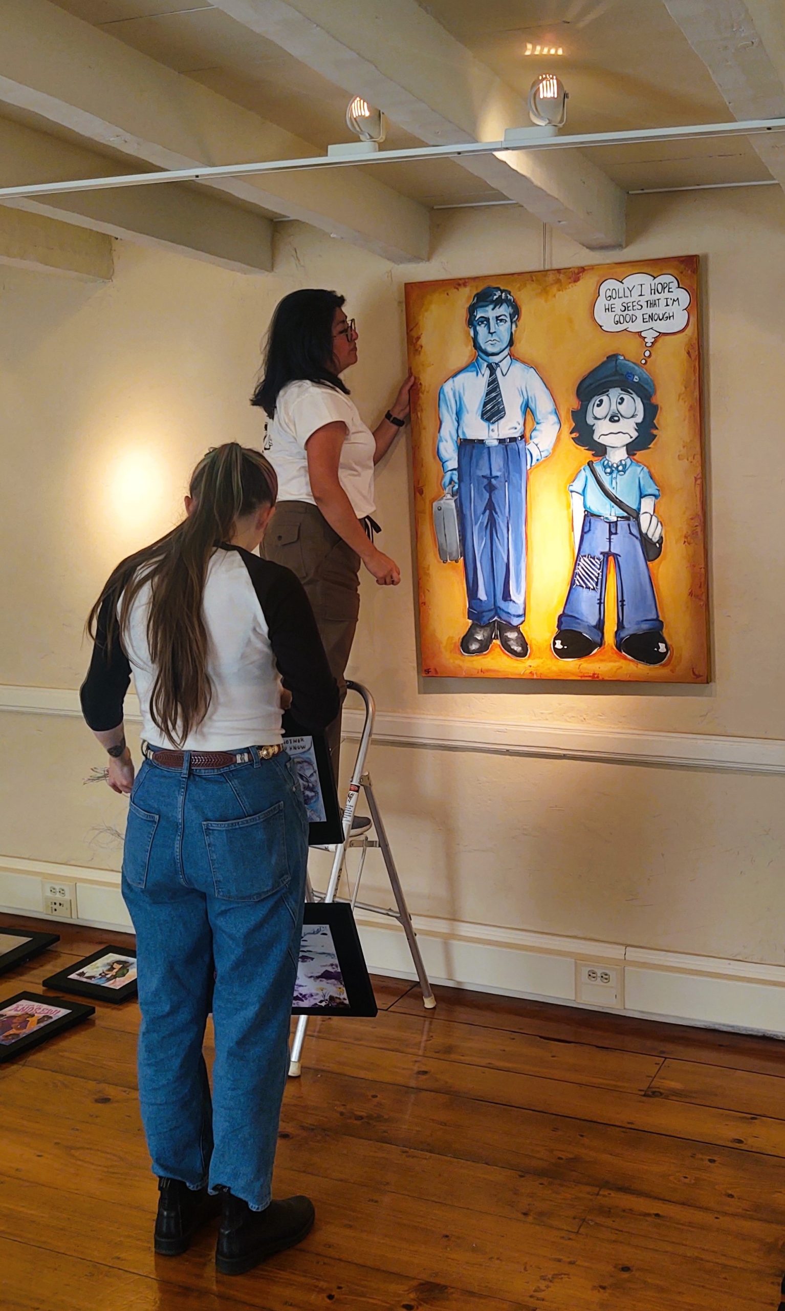 Two people stand in a well-lit art gallery space with warm wooden floors and white walls. To the right, a large, rectangular painting hangs prominently. The painting, rendered in a colorful, cartoonish style, shows two figures: one tall, dressed in a blue shirt, tie, and pants, holding a black briefcase; the other shorter, wearing a blue hat, shirt, and overalls, carrying a brown satchel. A speech bubble next to the shorter figure says, "Golly I hope he sees that I'm good enough". One person is on a stepladder, adjusting the artwork while the other stands at the foot of the step ladder. Several framed artworks are visible on the floor in the foreground.
