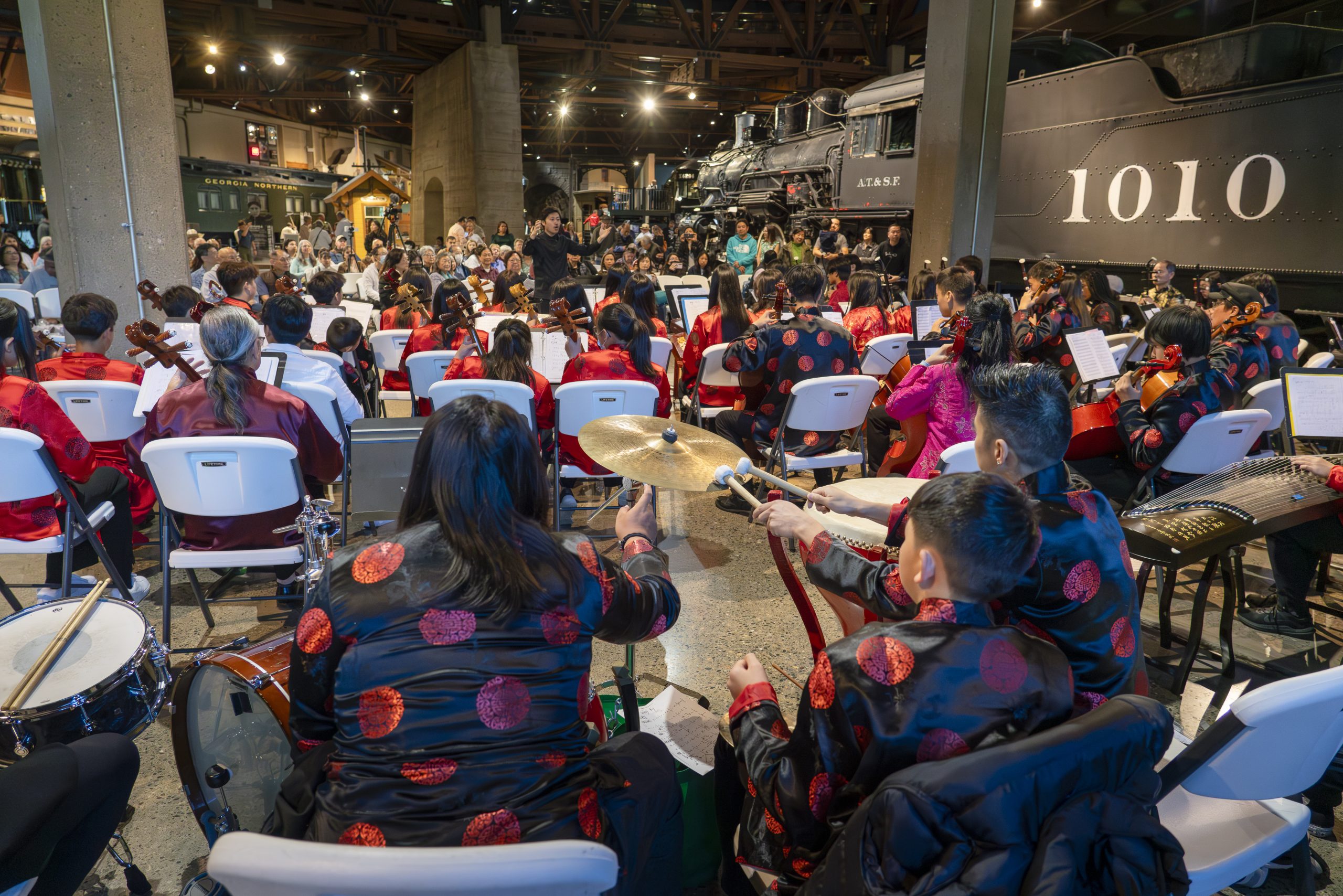 Three youth orchestra members sit with their backs facing the camera, wearing black silk shirts with red flowers. They are grouped around several percussion instruments. In front of them is the rest of the youth orchestra, mostly wearing red silk shirts with black embroidery, with a conductor standing and gesturing. In the background, a seated audience watches. The high-ceilinged museum room is ringed with full-size train cars.