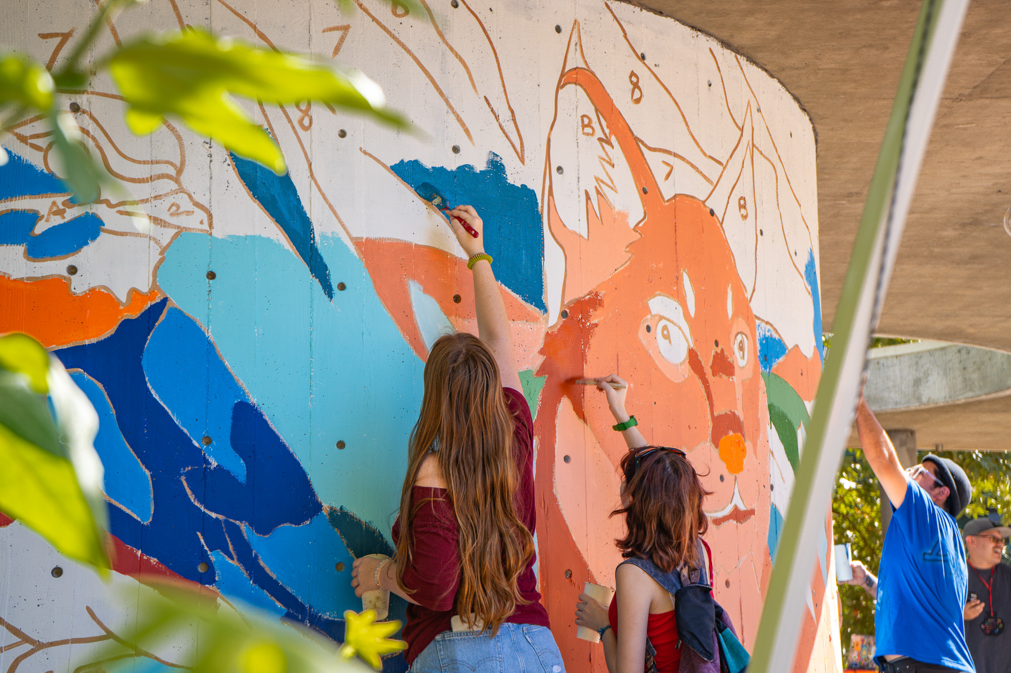 People painting a large mural on a concrete wall. Two people in the foreground, both with long hair, are adding vibrant colors to the mural, using brushes to apply blue, orange, and teal paint. The mural appears to depict a bobcat or an animal with large ears and expressive eyes. The wall has numbered sections to guide the painting process, like a large-scale paint-by-numbers artwork. In the background, two other individuals are engaged in the painting activity. Bright green leaves in the foreground frame the scene, adding a natural touch to the lively artistic environment.