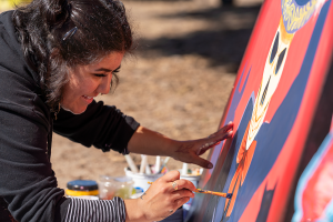 A smiling dark-skinned woman grins while painting a large mural in bright colors. She's bracing one paint-covered hand against the mural board while painting with the other. Everything is sunny and warm.
