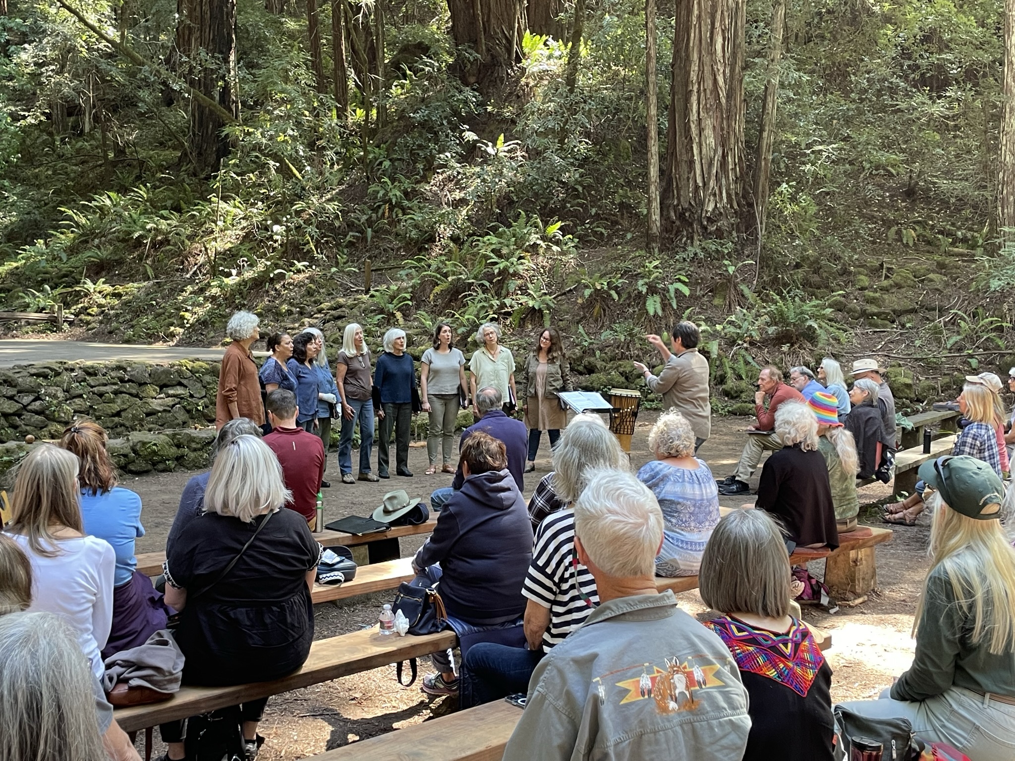 Group of 9 standing singers are led by a conductor, in front of an audience of around 150 people, against a backdrop of redwoods and ferns.
