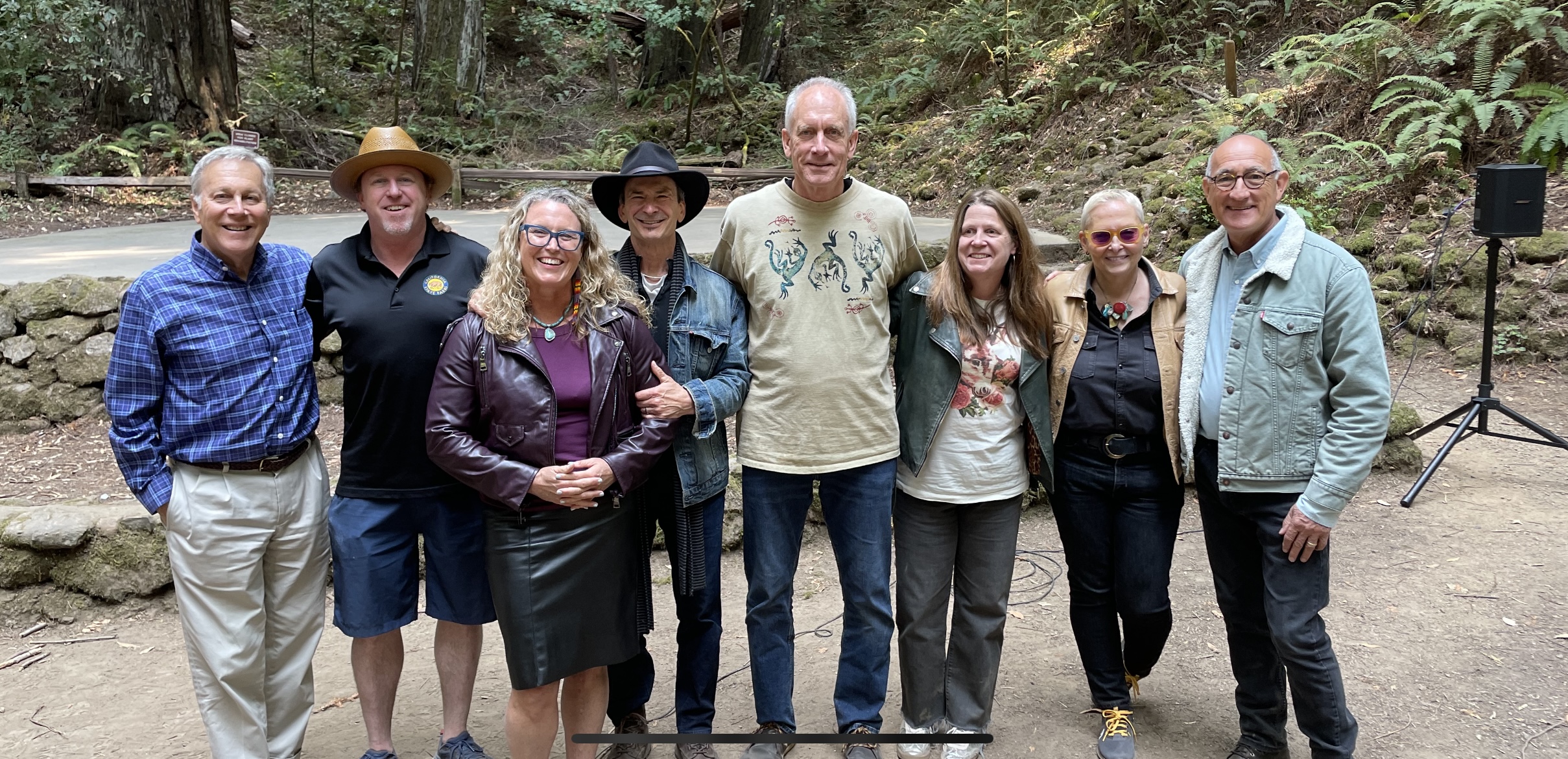 A group of seven people standing together in an outdoor forest setting, smiling at the camera. They are dressed in casual and comfortable attire, with trees and greenery in the background, suggesting a nature setting or park. The group appears happy and relaxed, enjoying their time together in the natural environment.