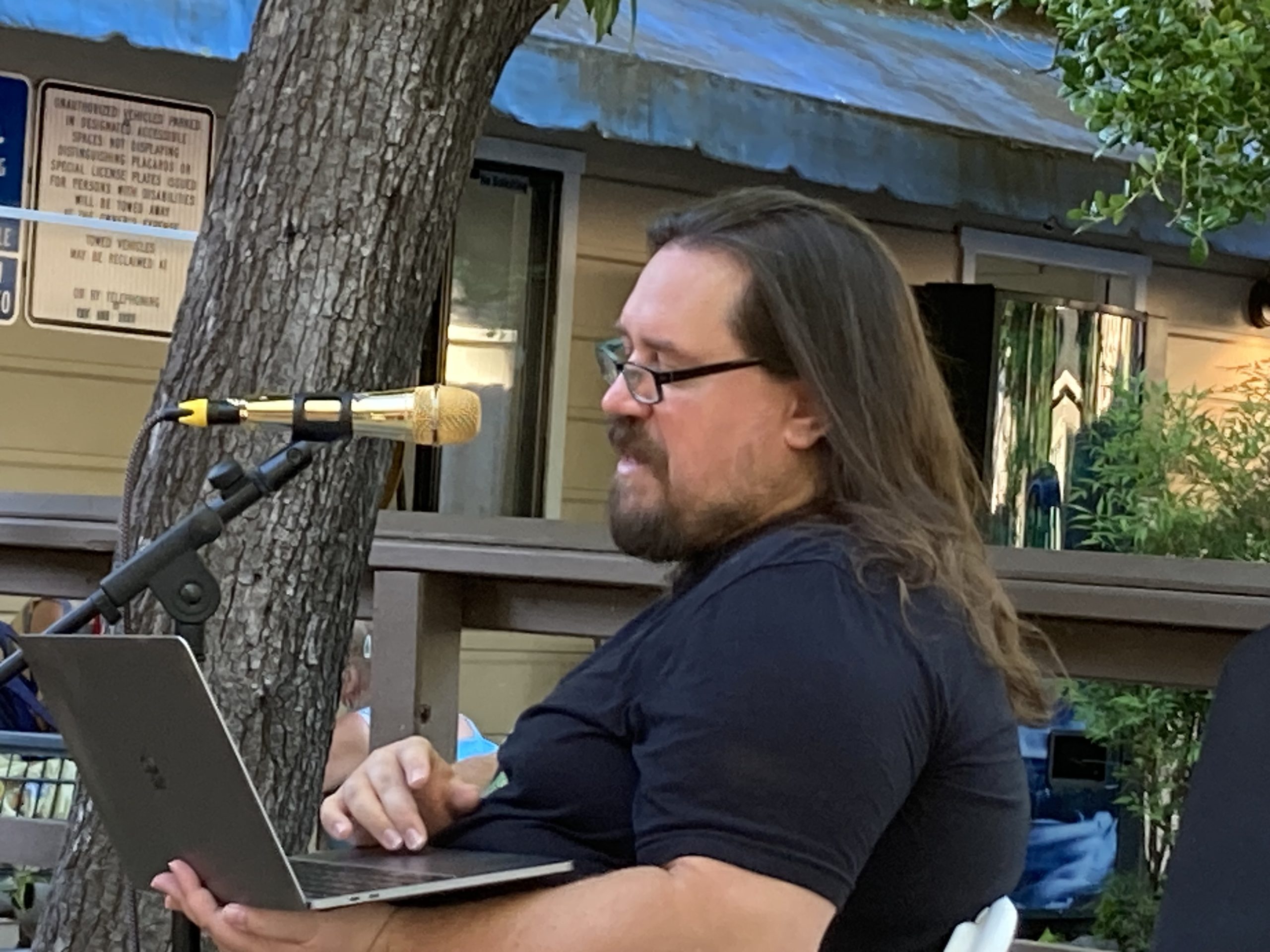 A man with long hair and glasses sits and speaks into a microphone as he reads off a laptop. He is wearing a black shirt, and there is greenery and a building in the background.