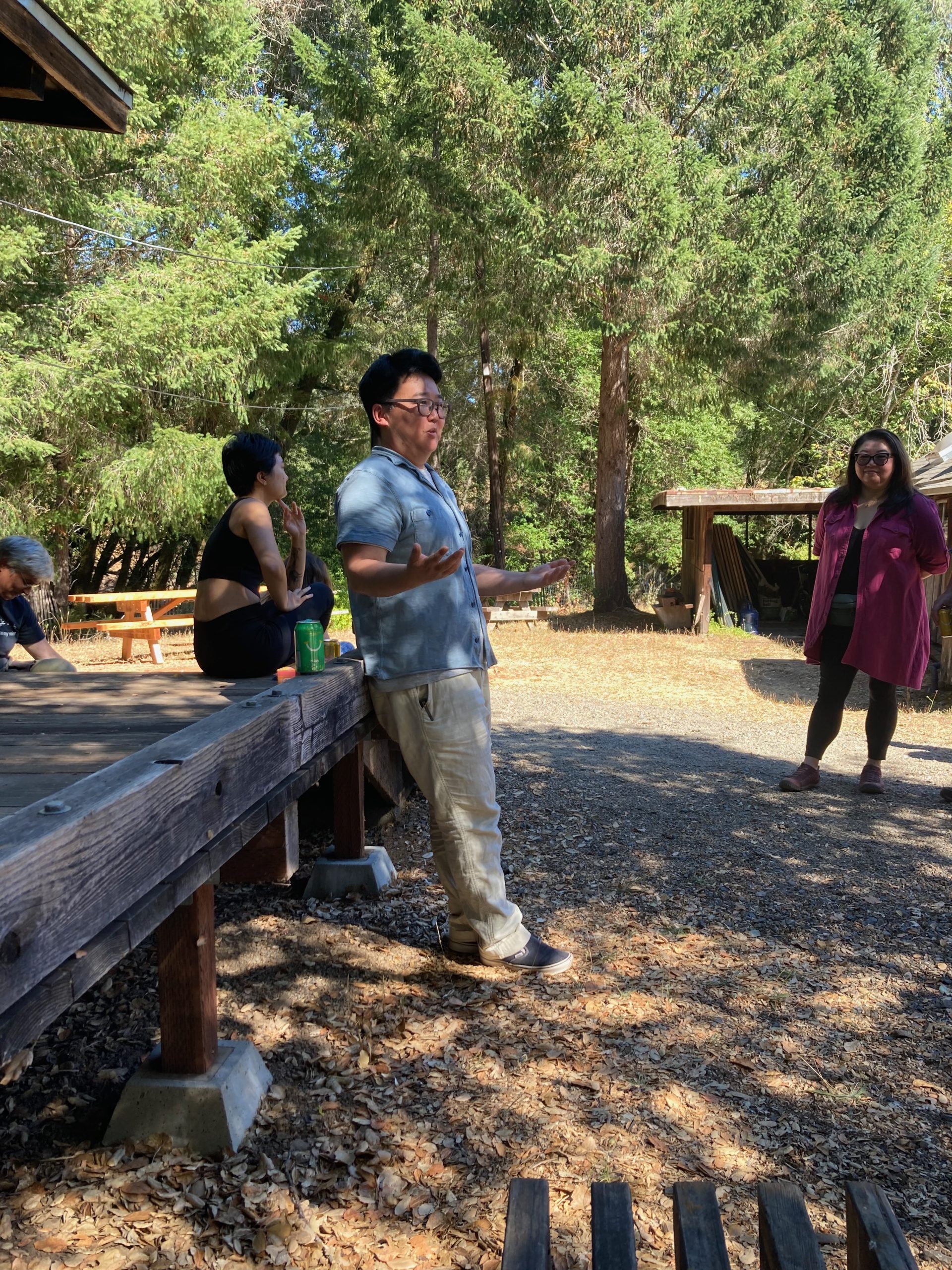 People are gathered outdoors in a wooded area, standing or sitting near a wooden platform. The focus is on a person wearing a light blue shirt and glasses, gesturing as they speak, with two other individuals listening attentively. The background features tall green trees, wooden structures, and scattered leaves on the ground, creating a serene natural setting.