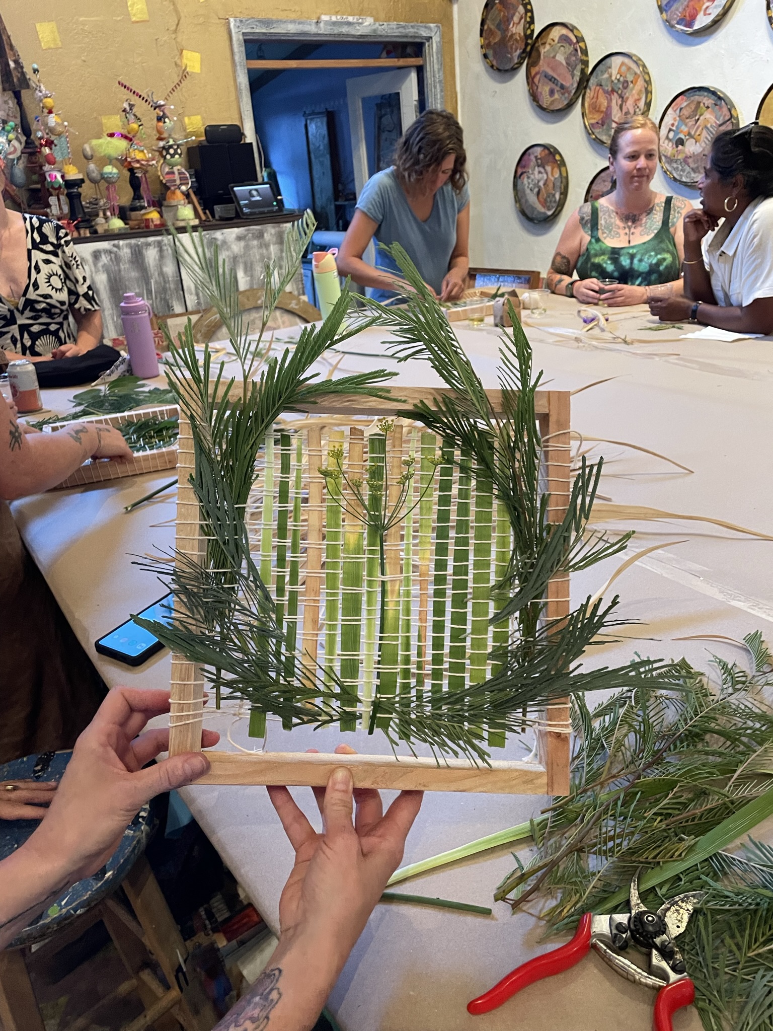 A handcrafted artwork made from natural materials, primarily featuring a woven structure framed by a wooden square. The woven portion consists of thin strips of green and beige leaves or reeds, interlaced vertically and horizontally. Fresh green foliage is woven around the edges of the frame, creating a textured border. In the background, people are engaged in similar activities, and vibrant, colorful artworks are displayed on the walls. The setting appears to be a workshop or art studio focused on creative and sustainable crafts.