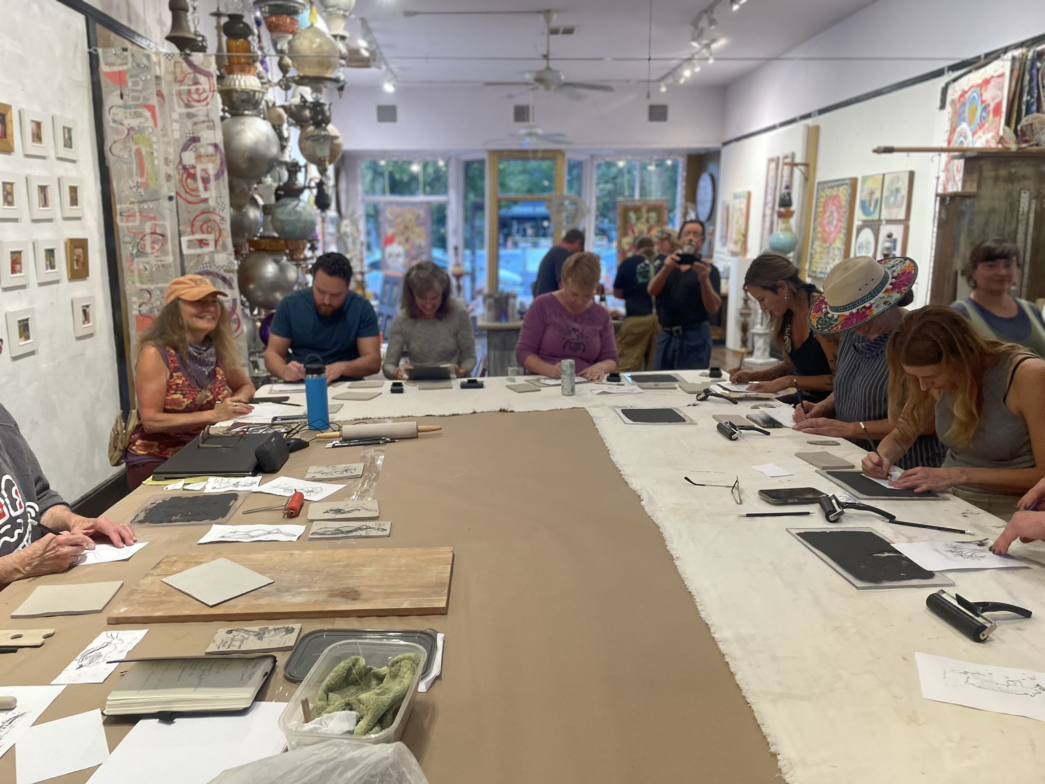 A group of people gathered around a large table in an art-filled studio, working on a ceramic monoprint project. The table is covered with tools like rollers and sketchbooks, and small artwork pieces. The room has colorful, eclectic decor, including paintings on the walls and an array of metallic lamps hanging from the ceiling. The atmosphere is lively and creative, with participants fully engaged in their artwork.