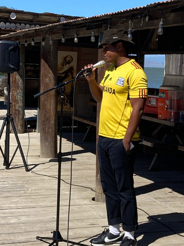 A person standing on a wooden platform, speaking into a microphone that is mounted on a stand, wearing a yellow Uganda national team jersey, black pants, white socks, black and white athletic shoes, and a dark wide-brimmed hat. The person has one hand in their pocket while delivering his speech. The background features a rustic wooden structure with string lights, a poster, and a cooler. A glimpse of the ocean can be seen in the distance, indicating an outdoor setting by the water.