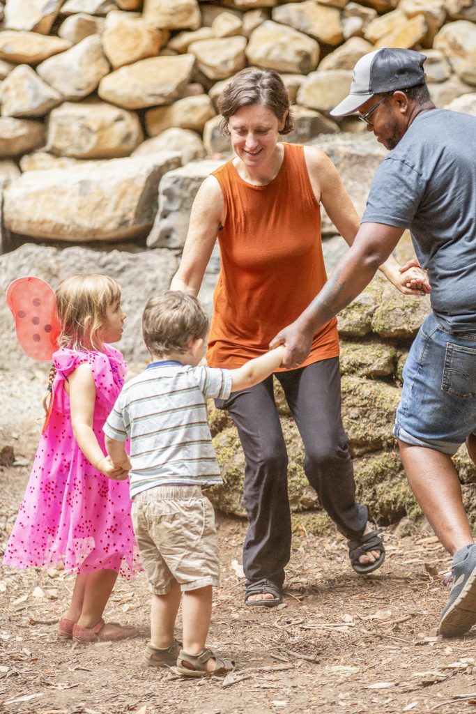 Two adults, one wearing an orange sleeveless top and the other in a gray T-shirt and cap, are holding hands with two young children. One is dressed in a pink dress with wings, and the other in a striped shirt and beige shorts. All holding hands in a circle, standing on a dirt path, with a backdrop of large, rustic stones. The group appears to be engaged in a playful activity, creating a scene of warmth and togetherness.