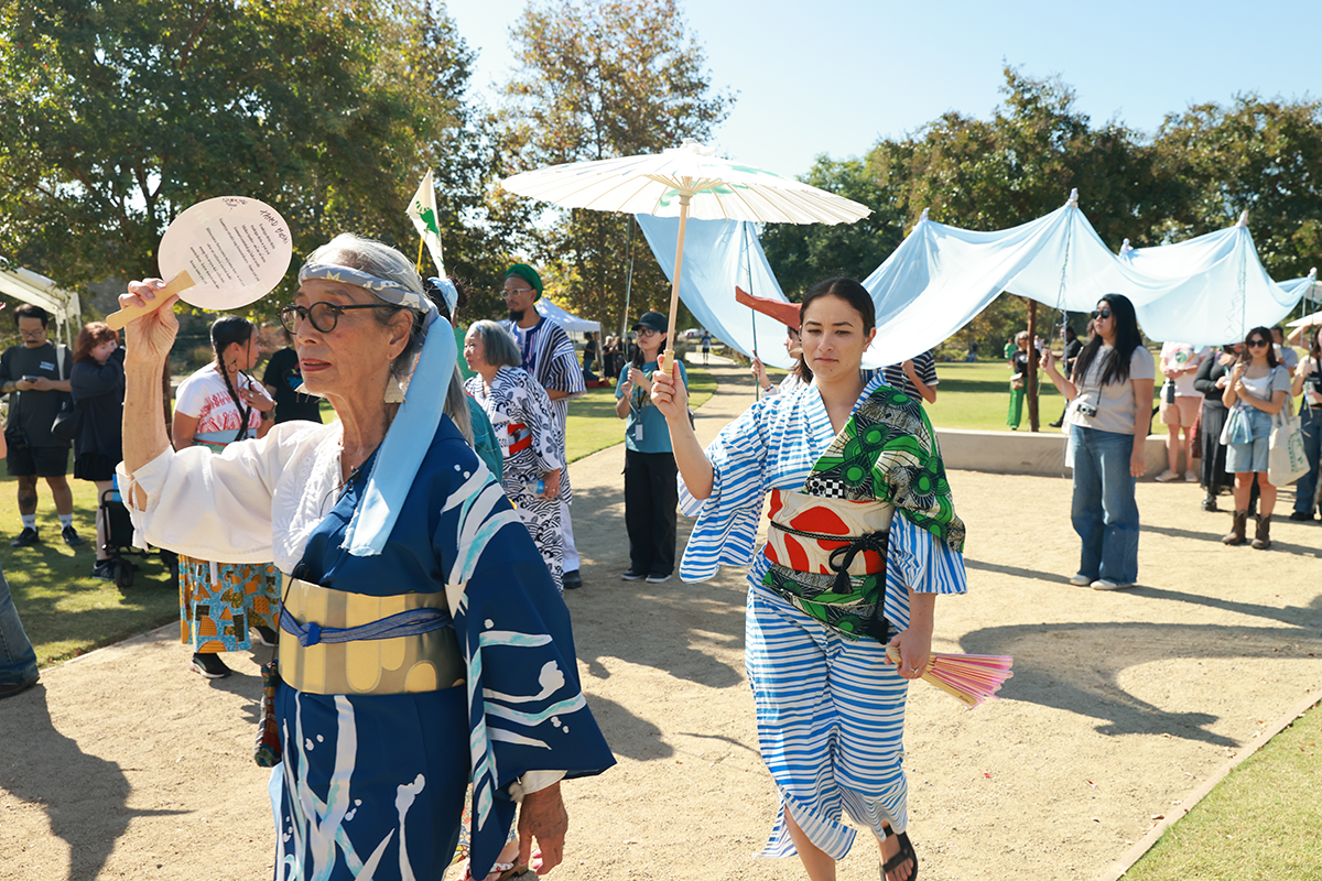 A bright sunny day along a gravel path in a park. The path is full of poeple dressed in traditional Japanese robes, most of whom are also carrying paper parasols and other shade. In the background are several folks holding support poles for a light blue ribbon of fabric representing the Los Angeles River.