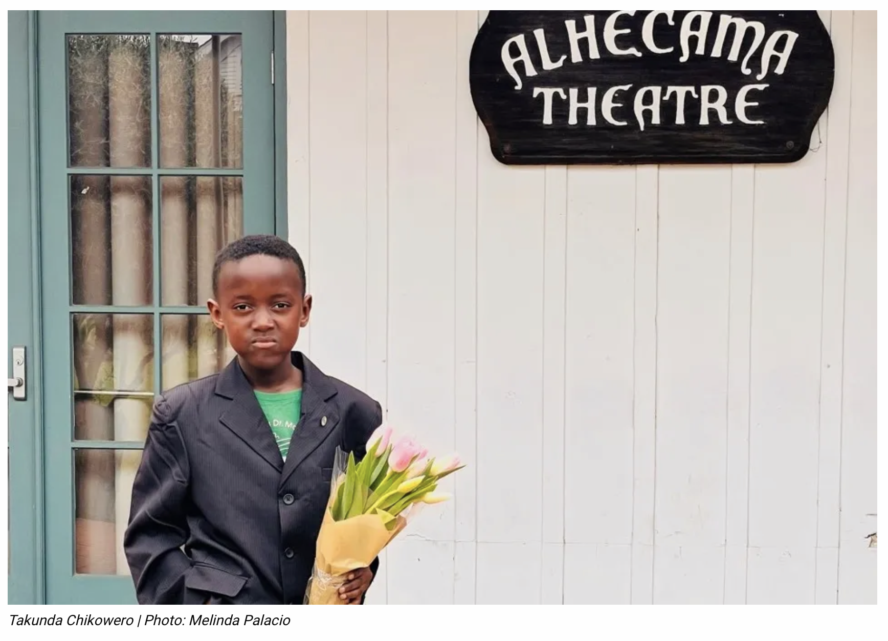A young Black boy wearing a sport jacket and holding a bouquet of pink tulips stands with one hand in his pocket against the outside of a building next to a black wooden “Alhecama Theatre” sign. He is looking at the camera with a serious and proud expression.