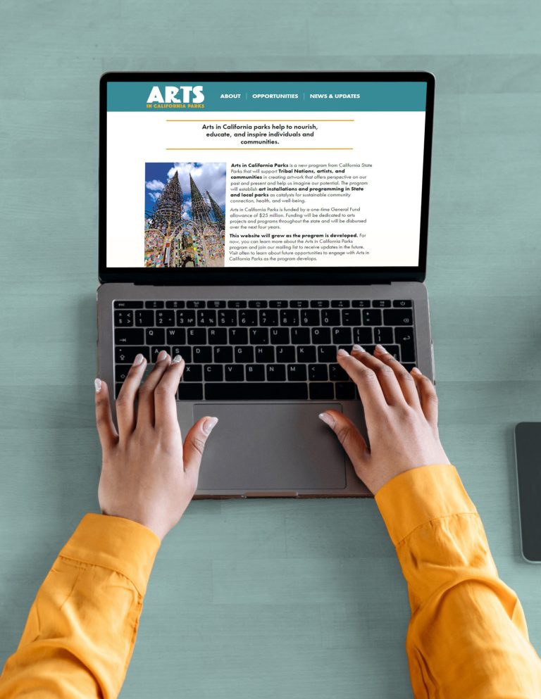 A laptop showing this Arts in California Parks site's homepage on a green table from the point of view of the person using it. Two arms in yellow sleeves reach forward towards it, brown hands poised over the keyboard.