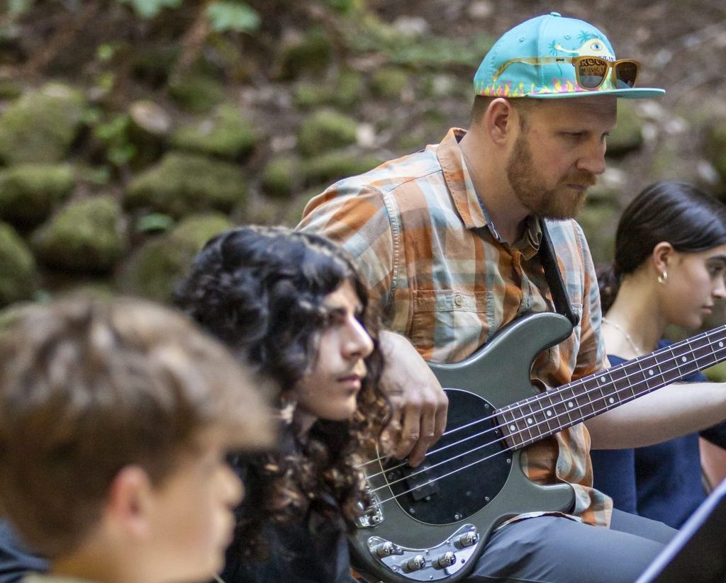 Several musicians perform outdoors in a lush, forested area. The bassist, wearing a plaid shirt and a turquoise cap, is focused on his instrument and the performance.